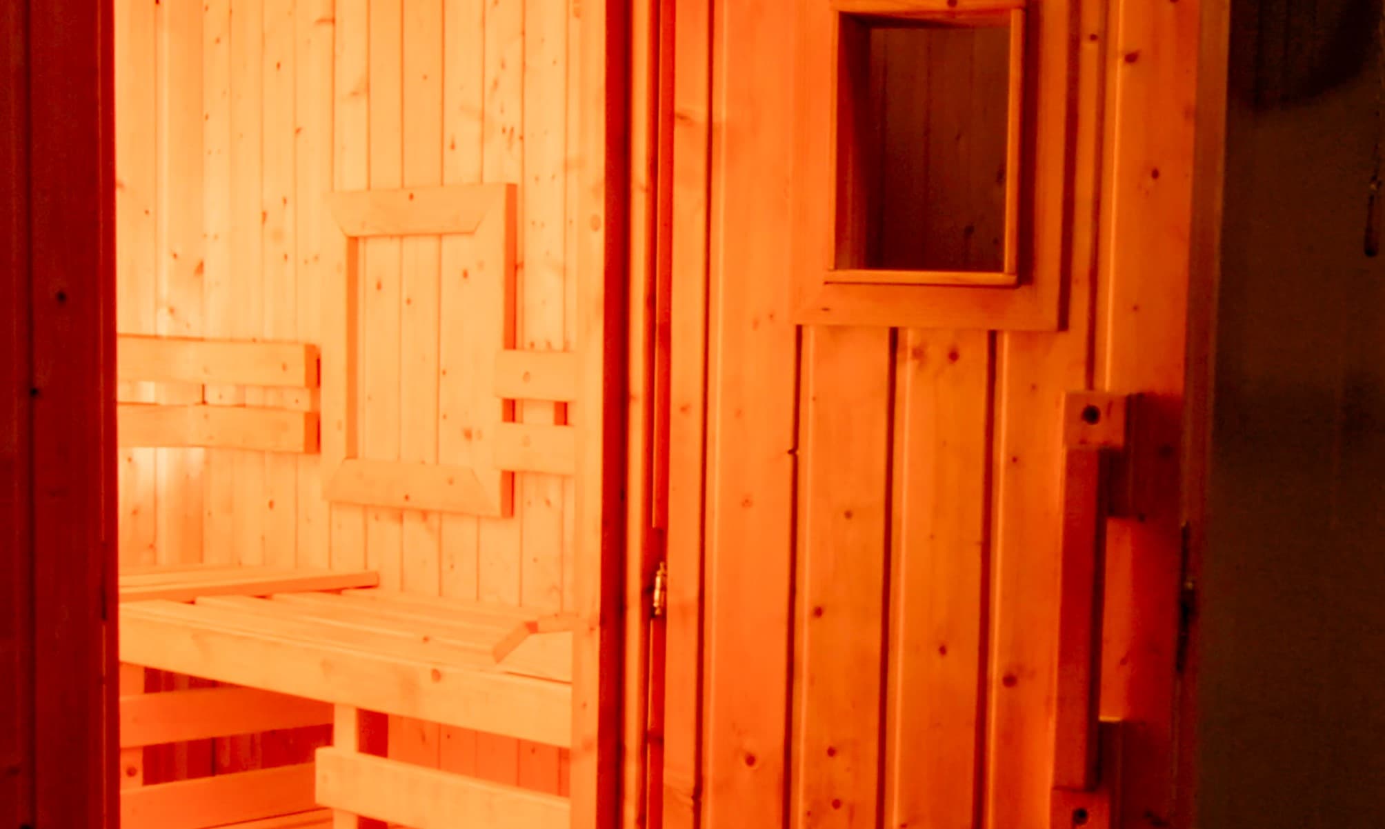 Das Innere einer Sauna mit einer offenen Tür, die Holzbänke und Paneele mit rotem Licht hat, was eine ruhige Atmosphäre verleiht.
